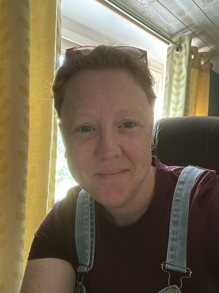 Author Picture Author portrait of a smiling person wearing a maroon shirt and denim suspenders, seated near a window with yellow curtains, soft natural lighting